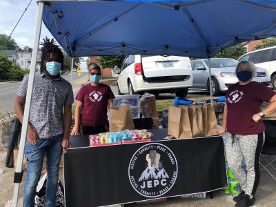 Three people wearing masks standing beneath a pop-up awning with a table of supplies