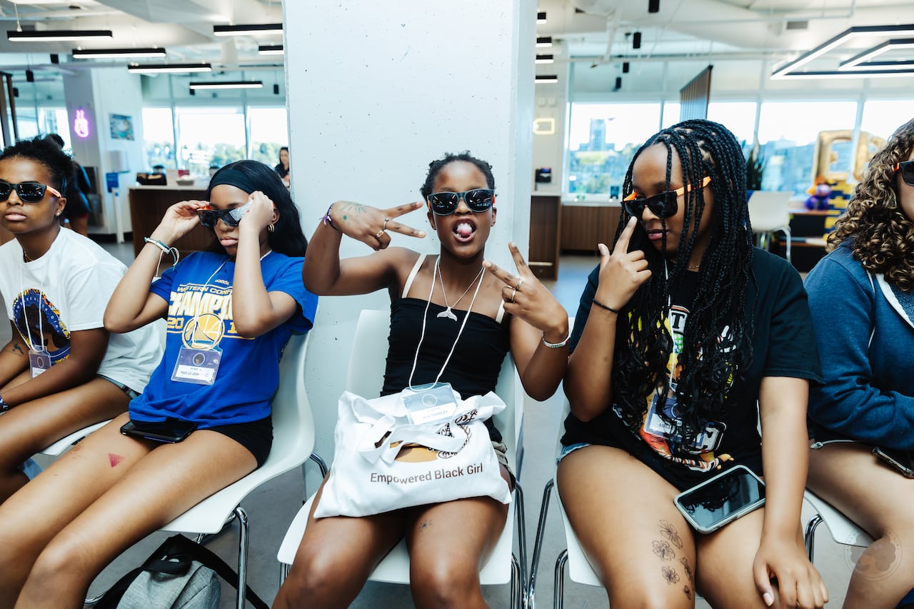 Five young girls wear sunglasses. The one in the middle is showing attitude with the V sign palm inward on each hand.