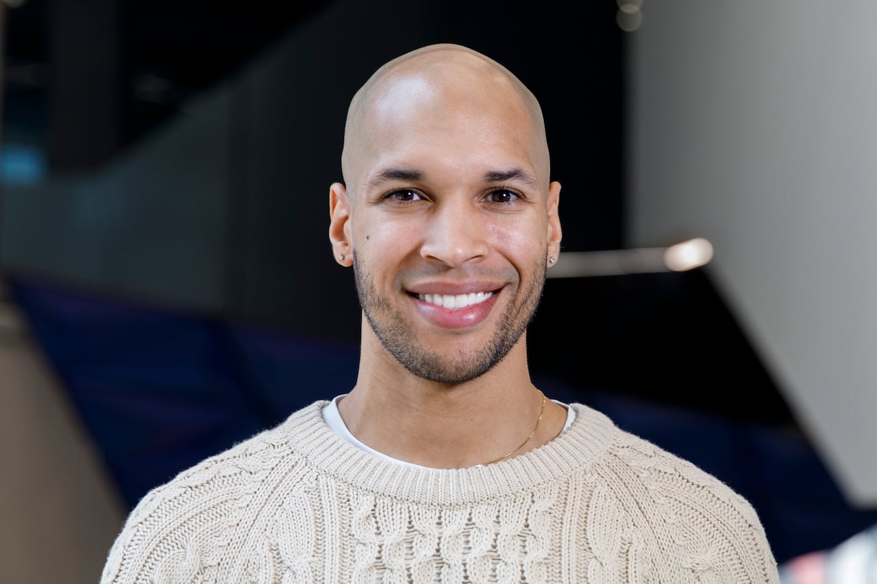 A Black man wearing a cream coloured sweater, thin gold necklace and diamond stud earrings smiles at the camera. 